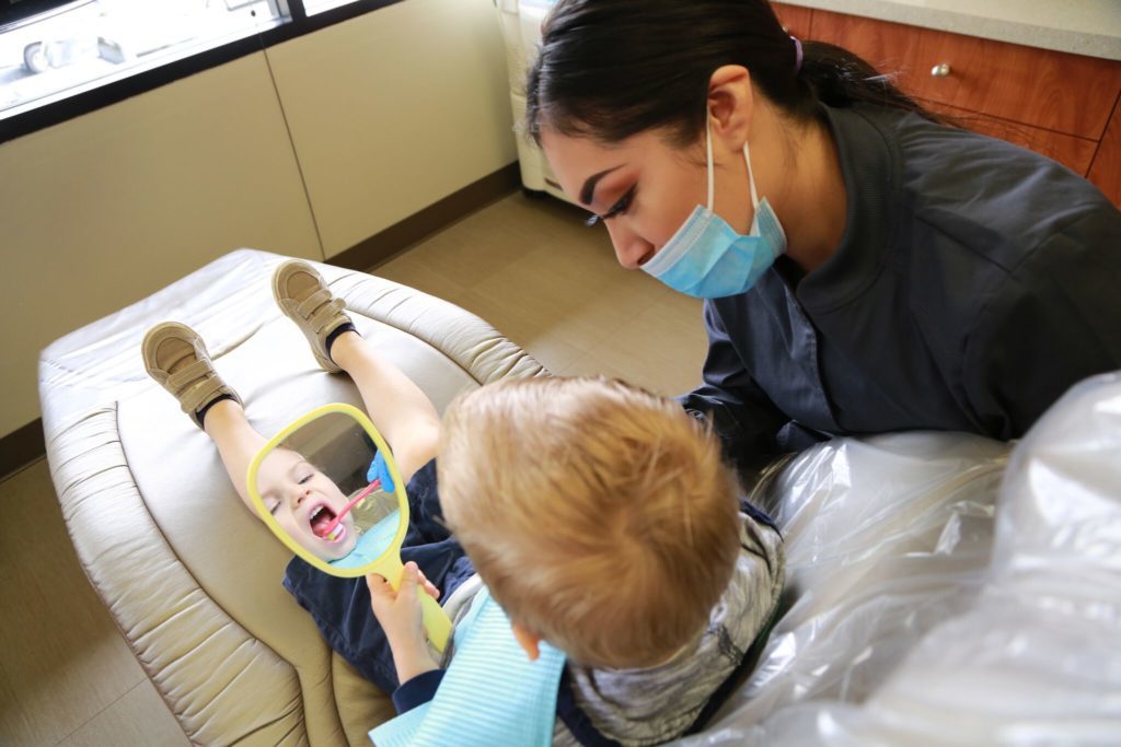 A young pediatric dentistry patient holds a mirror as the hygienist show him how to brush his teeth.